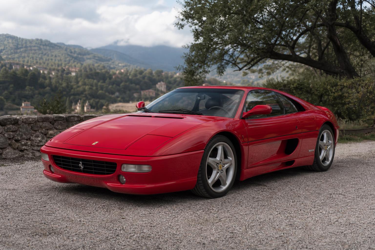Red Ferrari F355 Berlinetta parked on a gravel path with a scenic backdrop of rolling green hills and a stone wall under a cloudy sky.