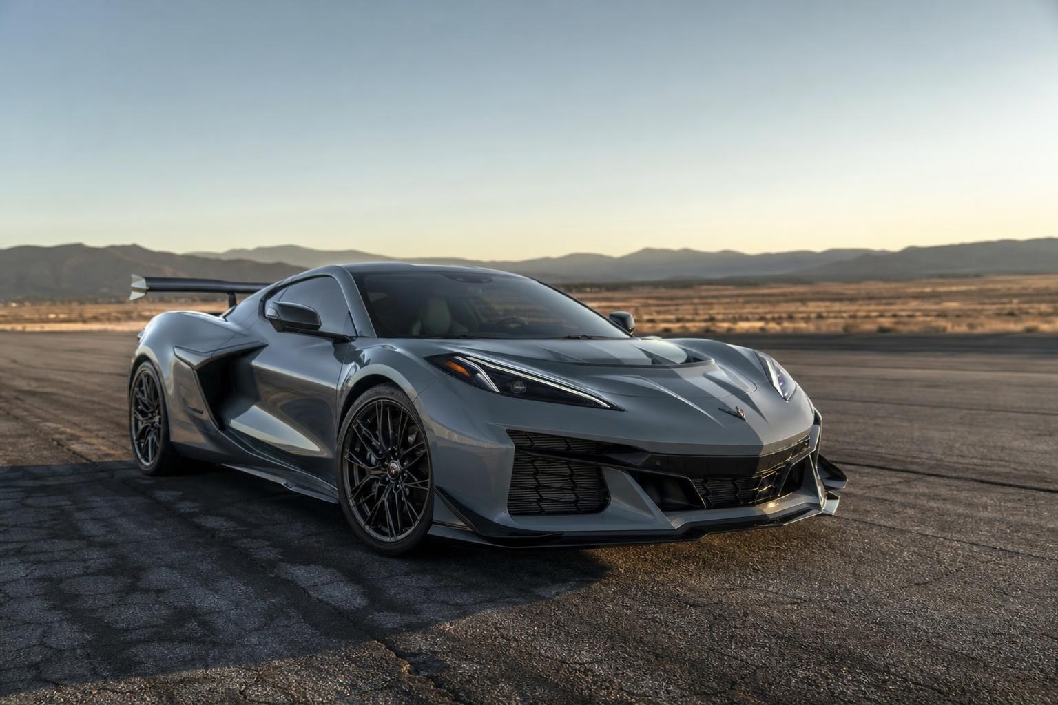 Corvette ZR1X 2026: Potencia e Tecnologia Sleek grey Corvette ZR1X with a large rear wing parked on an asphalt track against a desert mountain landscape at sunset.