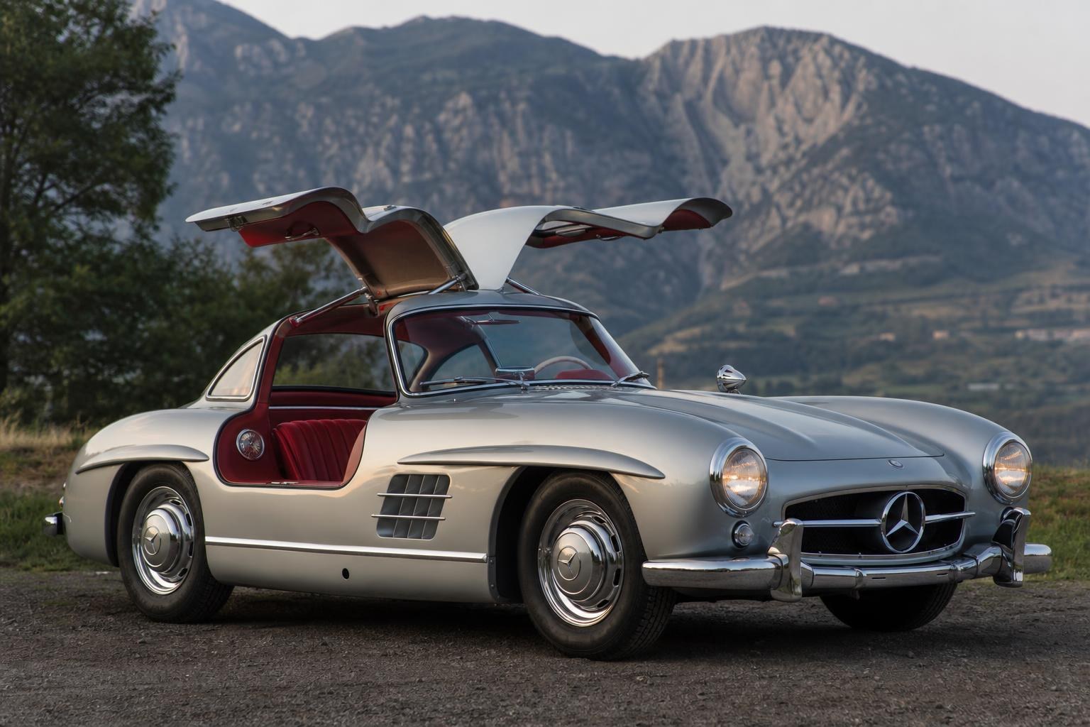 Mercedes-Benz 300SL Gullwing: A História do Ícone que Mudou o Automobilismo Silver Mercedes-Benz 300SL Gullwing with its signature doors open, featuring a red leather interior and parked against a rugged mountain backdrop.