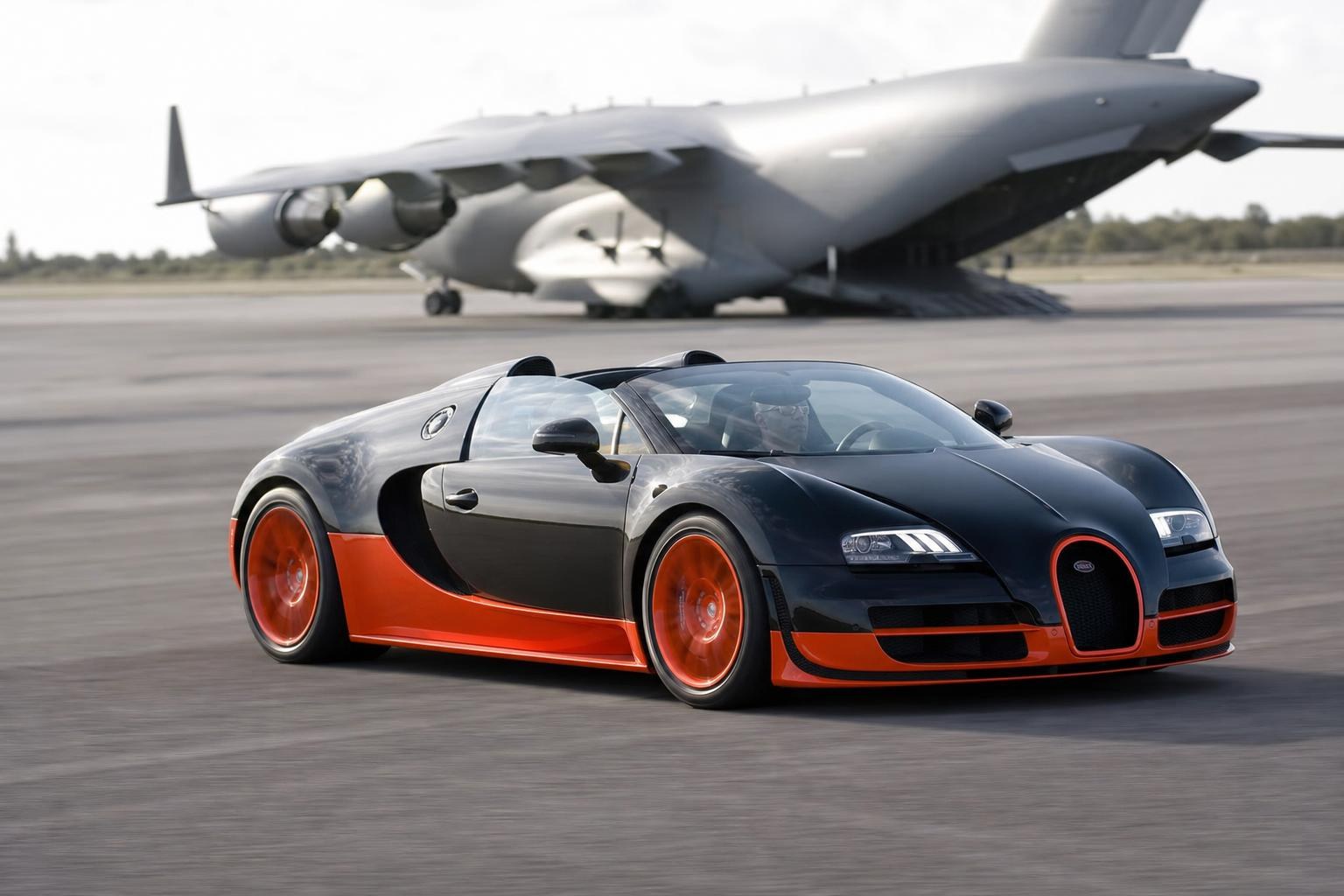 Black and orange Bugatti Veyron Grand Sport Vitesse driving on a runway with a large grey military cargo plane in the background.