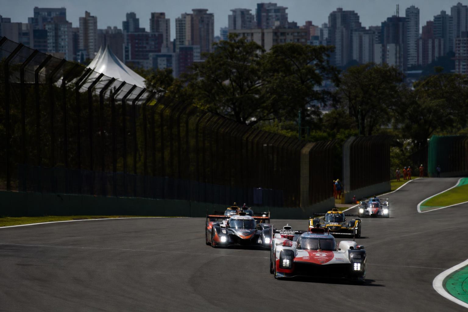 Endurance race cars speeding around a curved track at Interlagos with the São Paulo city skyline and trees in the background.