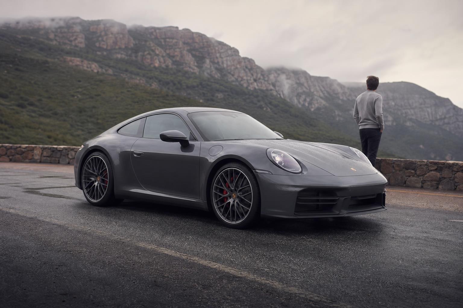 Porsche 911 (992.2): Evolução de um Clássico Grey Porsche 911 Carrera parked on a wet mountain road under a cloudy sky, with a man standing by a stone wall looking at the misty peaks.